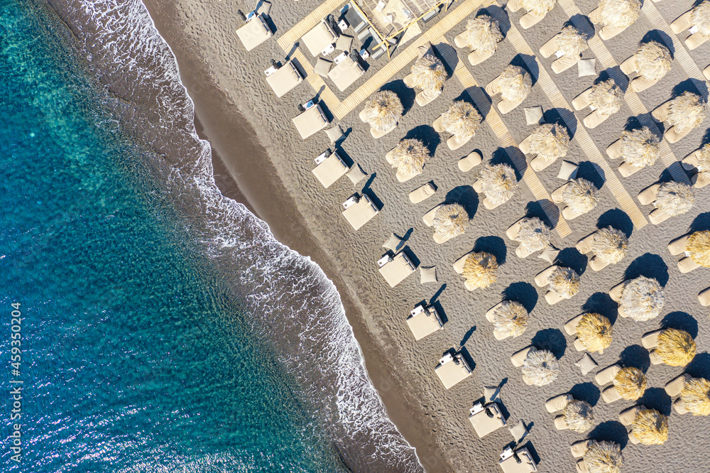 Top view aerial drone photo of black Perissa beach with beautiful turquoise water, sea waves and straw umbrellas. Vacation travel background. Aegean sea, Santorini Island, Greece