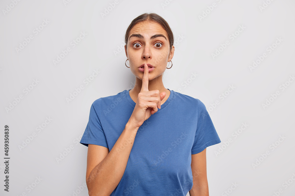 Surprised dark haired woman makes silence gesture presses index finger over lips dressed in casual blue t shirt asks keep quiet makes taboo sign poses against white background gossips about something Surprised dark haired woman makes silence gesture presses index finger over lips dressed in casual blue t shirt asks keep quiet makes taboo sign poses against white background gossips about something