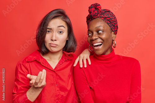Photo of mixed race diverse women react differently on something stand closely to each other against bright red background. Indignant Asian female says so what while her companion laughs joyfully