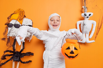 Glad small kid pretends being ghost or mummy holds halloween carved pumpkin stands against orange background poses near spooky toys says boo. Autumn holiday on 31st October. Scary special day