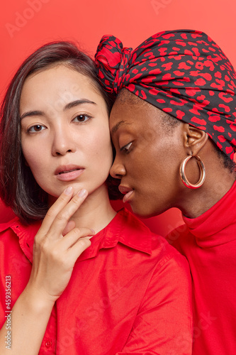 Serious multiethnic young women look at camera with confident expression dressed in casual clothes. Dark skinned female stands in profile wears silk scarf isolated over red background. Diversity