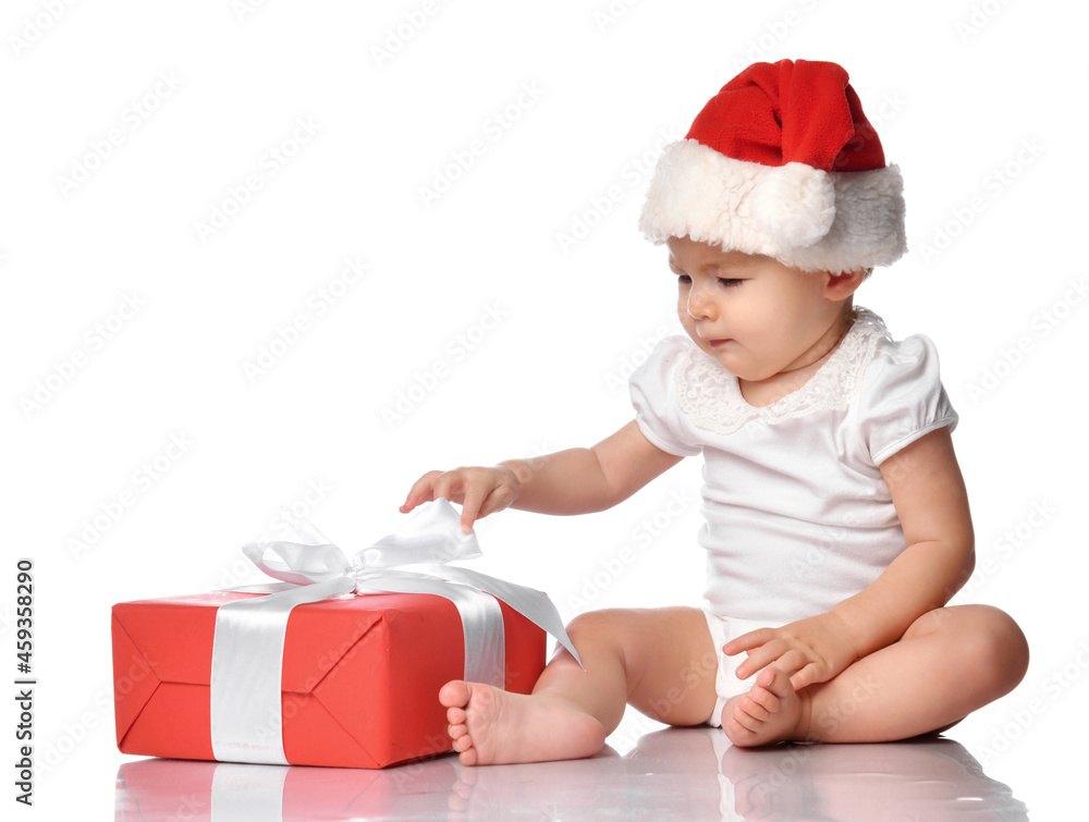 Cute infant baby girl wearing overalls, a red Santa Claus hat, sitting on a white floor studio, looking at a festive gift box wrapped with a bow. Isolated portrait. Christmas, New Year concept Cute infant baby girl wearing overalls, a red Santa Claus hat, sitting on a white floor studio, looking at a festive gift box wrapped with a bow. Isolated portrait. Christmas, New Year concept