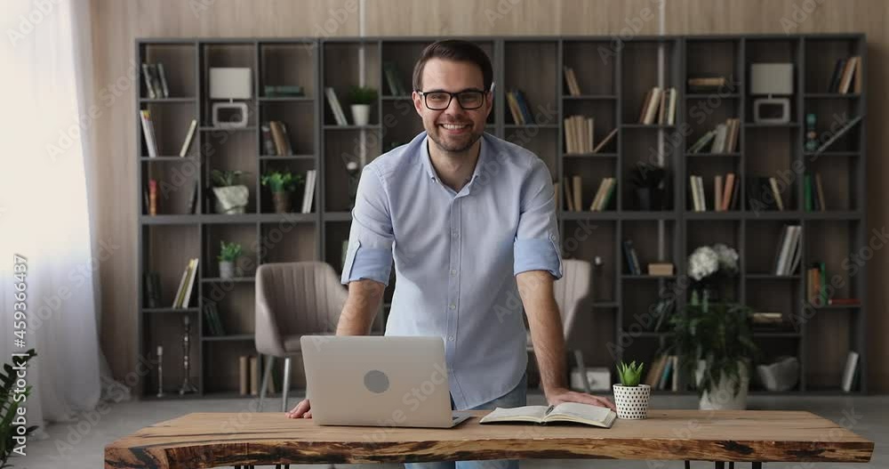 Smiling handsome confident young businessman standing at table with computer in modern office. Happy millennial generation male manager worker employee standing at workplace, successful career concept Smiling handsome confident young businessman standing at table with computer in modern office. Happy millennial generation male manager worker employee standing at workplace, successful career concept