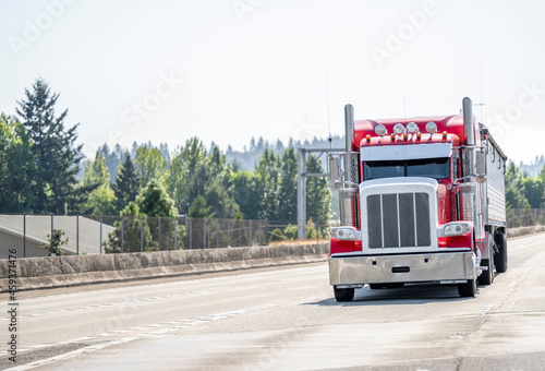 Bright red classic bonnet big rig semi truck with horns on the roof transporting cargo in covered bulk semi trailer running on the highway road in front of another traffic
