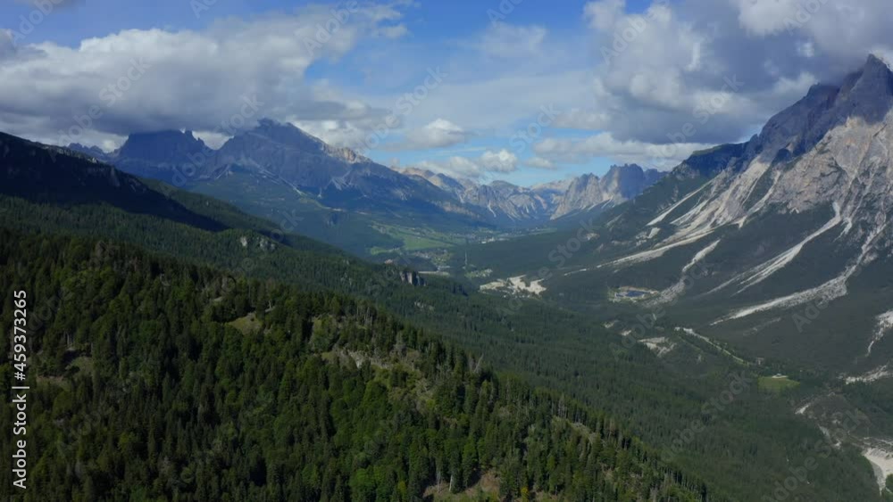 Dolomites, Cortina d'Ampezzo, Veneto, Italy, September 2021. Drone gradually tracks parallel with a forested hillside, into the populated valley surrounded by steep mountains reaching into the clouds. Dolomites, Cortina d'Ampezzo, Veneto, Italy, September 2021. Drone gradually tracks parallel with a forested hillside, into the populated valley surrounded by steep mountains reaching into the clouds.