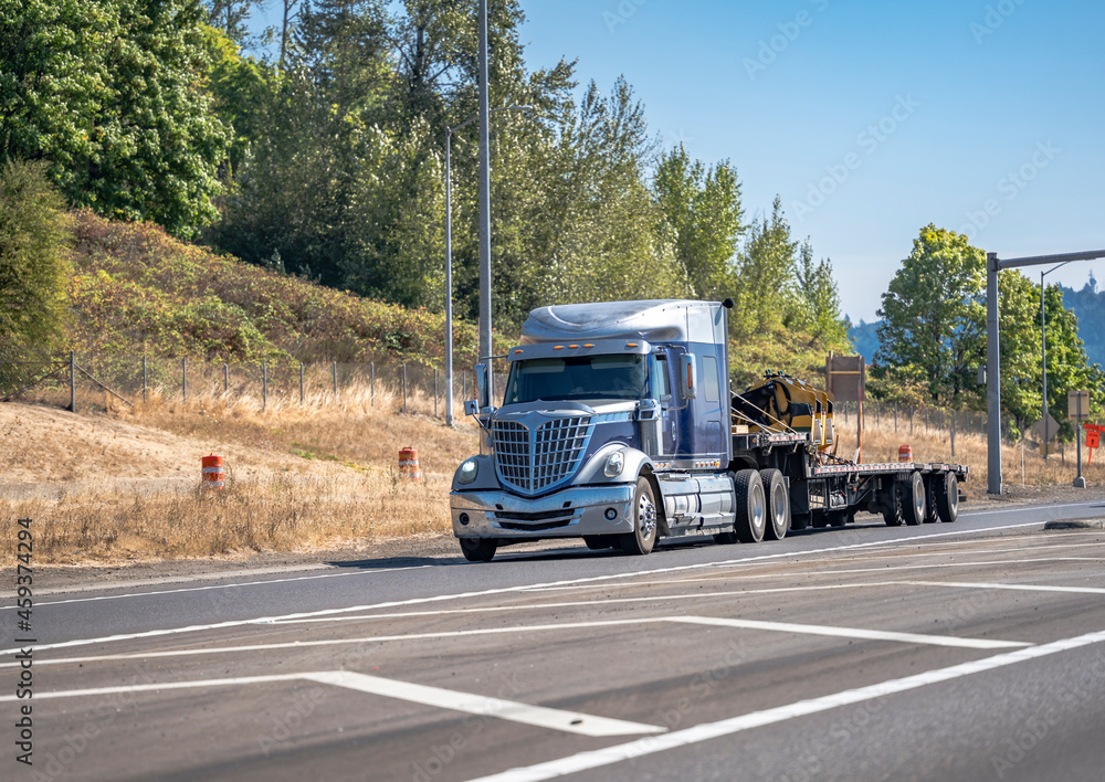 Stylish blue big rig bonnet star semi truck transporting construction equipment on step down semi trailer running on the highway road entrance.