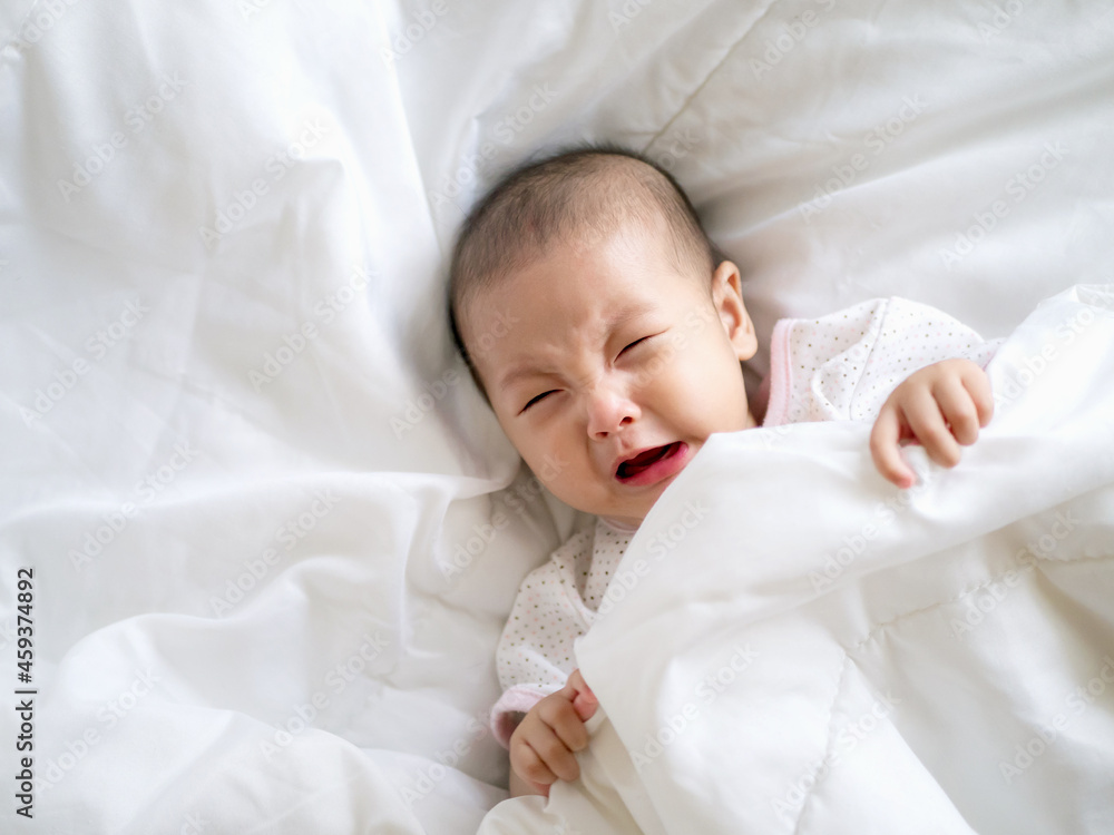 Close up Adorable Asian baby infant girl cry on a white bed , colic symptom or hungry unhappy of infant concept Close up Adorable Asian baby infant girl cry on a white bed , colic symptom or hungry unhappy of infant concept