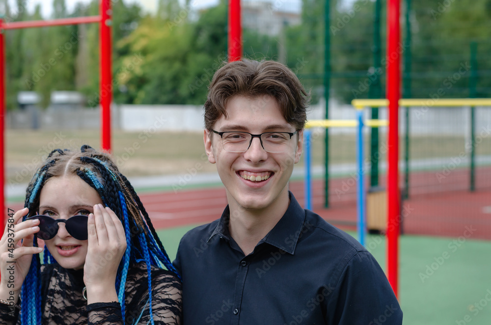 Obraz premium Laughing happy young woman and young man in front of a sports field. Heterosexual couple in love outside. Young adult date. Woman with blue pigtails. Man wearing optical glasses. Love, romance.