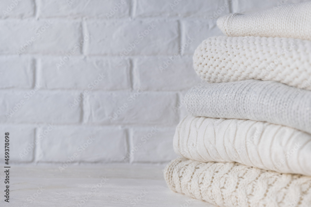 Stack of white cozy knitted sweaters against the background of white brick wall.