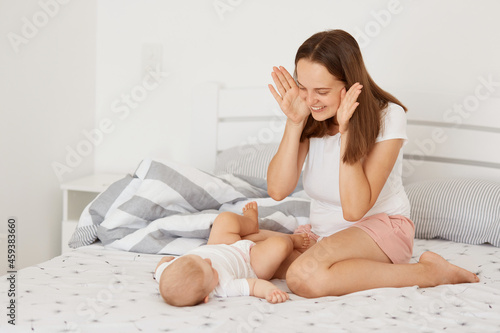 Portrait of happy young adult female wearing white casual t shirt sitting on bed and playing with her infant daughter peekaboo game, happy mother with toddle at home in light room.