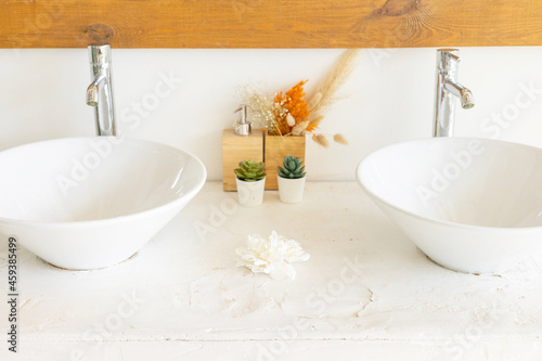 stylish and modern design bathroom interior with two white sinks  wicker baskets for cosmetics  mirror in wooden frame  dried flowers in vases  home plants in pot in Balinese style with white walls