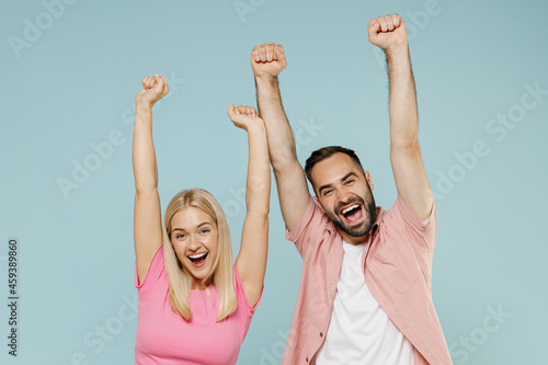 Young fun overjoyed excited couple two friends family man woman in casual clothes do winner gesture with raised up hands together isolated on pastel plain light blue color background studio portrait.