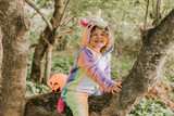 little girl in a rainbow unicorn costume kigurumi is sitting on a tree with pumpkin basket for sweets against the background of a forest. Halloween concept. space for text. High quality photo