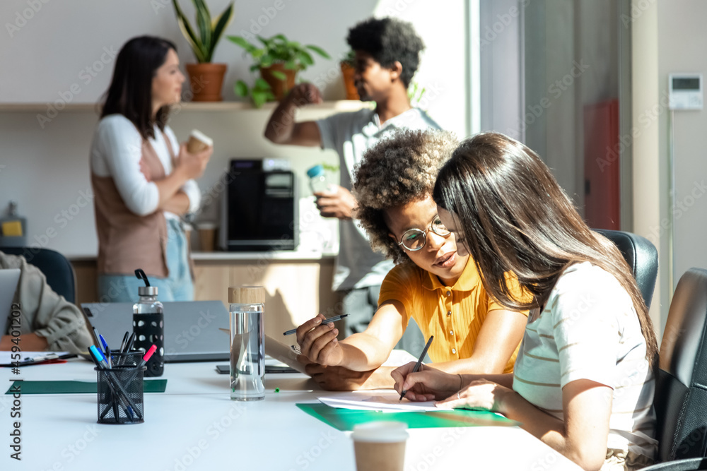 Two female coworkers share ideas while two colleagues have coffee break in the background Two female coworkers share ideas while two colleagues have coffee break in the background