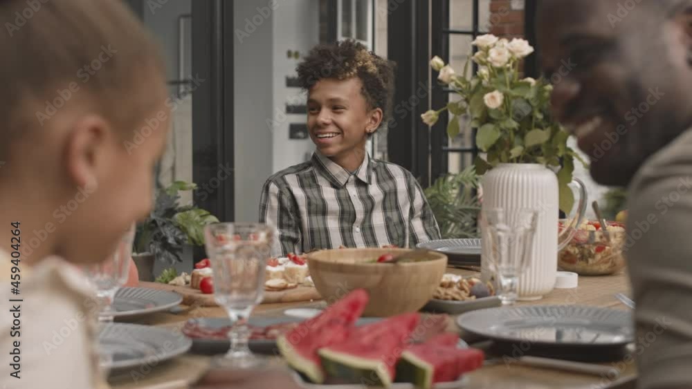 Medium close-up of teenage African boy smiling, talking to his family members sitting at dinner table on terrace at daytime