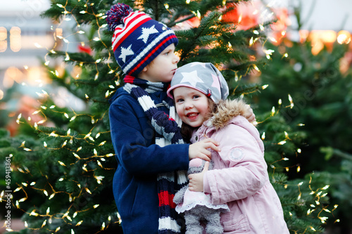 Two little smiling kids, preschool boy and girl hugging on German Christmas market. Happy siblings children in winter clothes with lights on background and xmas trees. Family funny brother and sister.