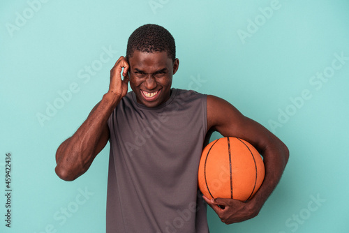 Young African American man playing basketball isolated on blue background covering ears with hands.
