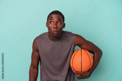 Young African American man playing basketball isolated on blue background shrugs shoulders and open eyes confused. © Asier