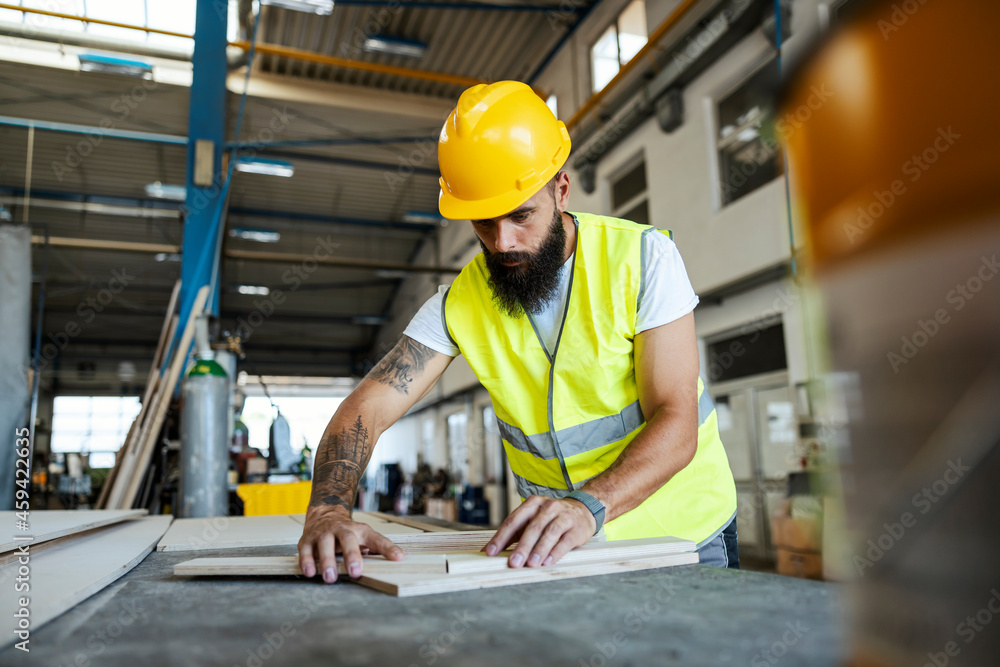 A bearded tattooed worker in a protective uniform is working with wooden parts in his workshop. Woodwork in workshop A bearded tattooed worker in a protective uniform is working with wooden parts in his workshop. Woodwork in workshop