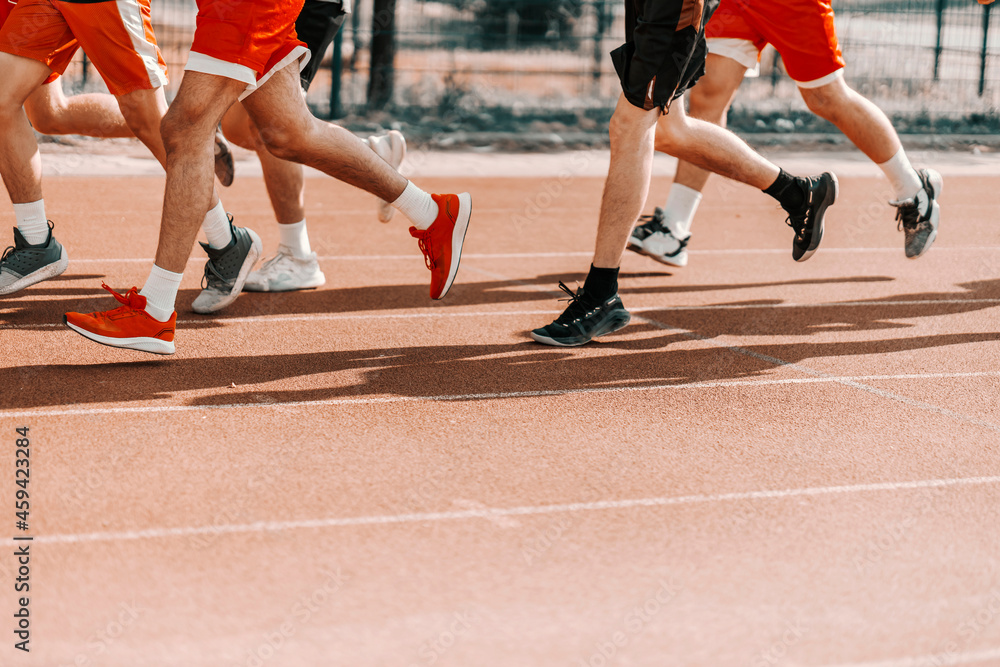 A group of runners at an athletic stadium. In the focus are the legs of a man's legs running on the track and warming up the muscles. Group fitness training and fitness marathon A group of runners at an athletic stadium. In the focus are the legs of a man's legs running on the track and warming up the muscles. Group fitness training and fitness marathon