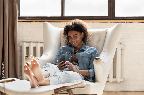 Smiling African American woman using smartphone, relaxing in armchair at home, happy attractive young female looking at phone screen, chatting, typing writing message in social network, browsing apps