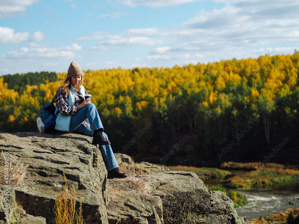 Naklejka premium Young curly woman traveller in plaid t-shirt sitting on a rock with coffee in her hands; there is mountain river on a background; sustainable tourism; ecological tourism; discovery and explore;