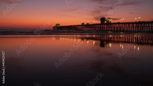 Pier silhouette Oceanside California USA. Pacific ocean tide tropical beach. Summertime gloaming atmosphere. Purple aesthetic gradient, calm twilight sky, pink violet dusk. Lights reflection in water. © Dogora Sun