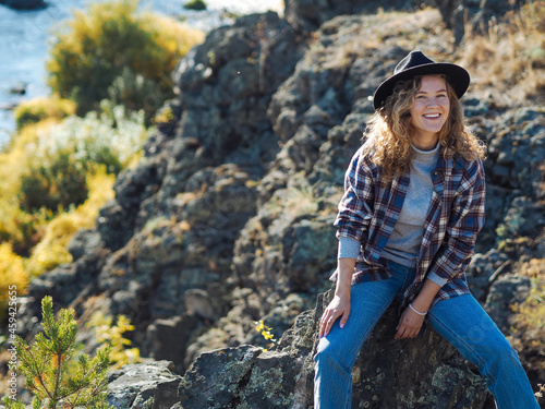 Young curly woman in hat and plaid t-shirt smiling on mountain hill with river view; autumn travel; beautiful nature; sustainable environment, eco tourism