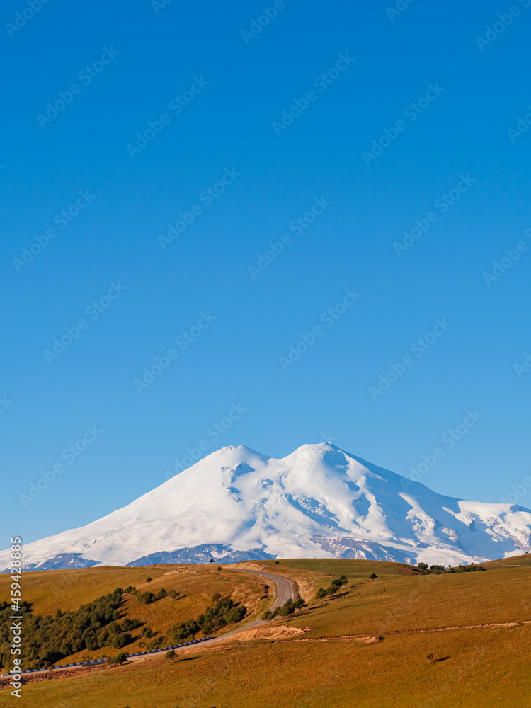 A beautiful asphalt road through the autumn hills of the Caucasus leads to Elbrus. Elbrus with two peaks against the blue sky is covered with snow. Copy space. A beautiful asphalt road through the autumn hills of the Caucasus leads to Elbrus. Elbrus with two peaks against the blue sky is covered with snow. Copy space.