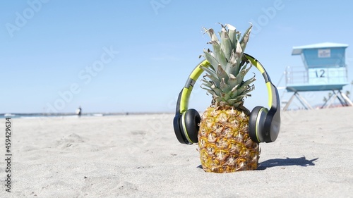 Funny pineapple in headphones, sandy ocean beach by blue lifeguard hut, California USA, Pacific coast. Tropical summer exotic fruit enjoying vacations and music in paradise. Ananas sunbathing on shore