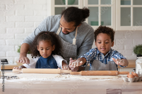Caring African American young father teaching little daughter and son rolling out dough, happy family spending leisure time in kitchen at home together, cooking baking homemade pastry or pie