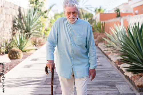 Suffering elderly man walking with the help of a cane. Senior white haired people outdoor in a tropical garden
