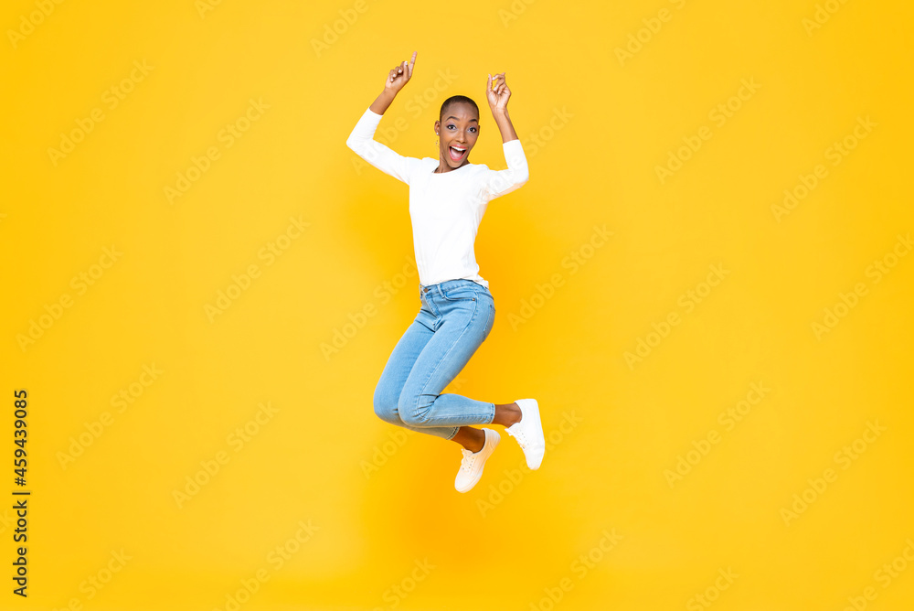 Energetic young African American woman jumping with hands pointing up in isolated yellow studio background