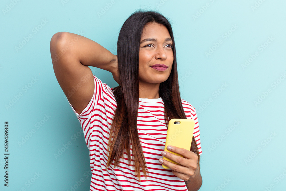 Young Venezuelan woman holding mobile phone isolated on blue background touching back of head, thinking and making a choice. Young Venezuelan woman holding mobile phone isolated on blue background touching back of head, thinking and making a choice.