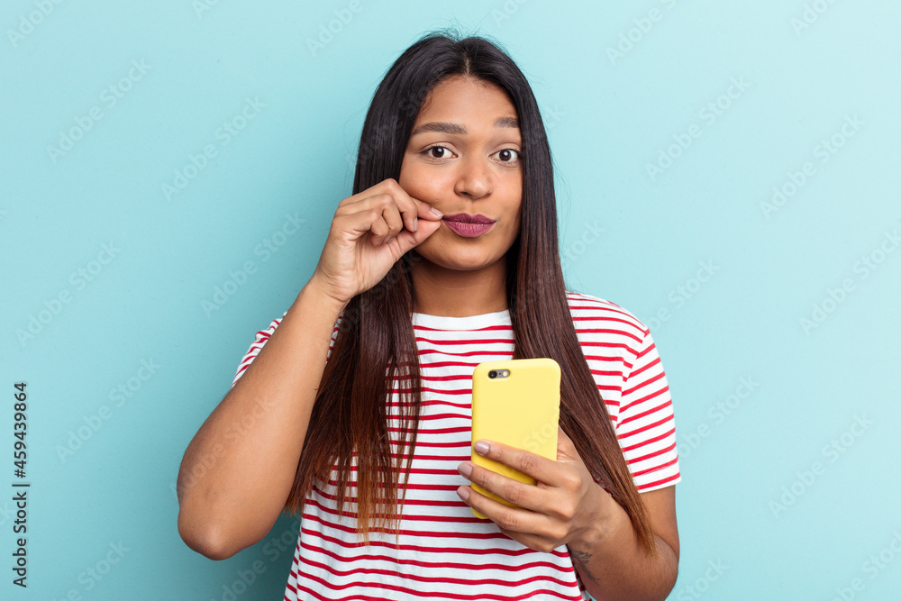 Young Venezuelan woman holding mobile phone isolated on blue background with fingers on lips keeping a secret. Young Venezuelan woman holding mobile phone isolated on blue background with fingers on lips keeping a secret.