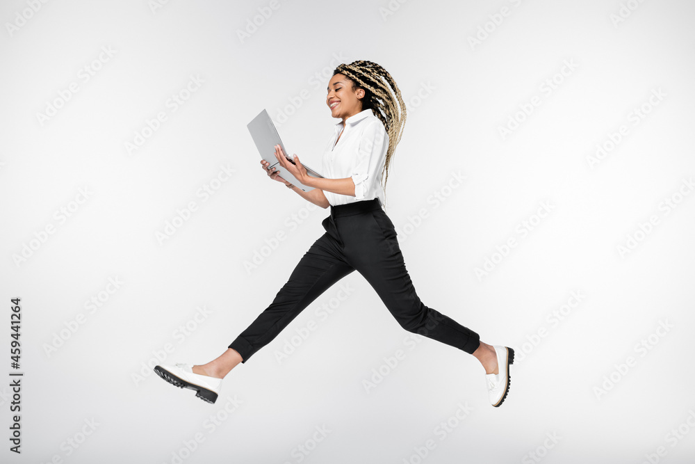 happy african american businesswoman levitating with laptop isolated on white