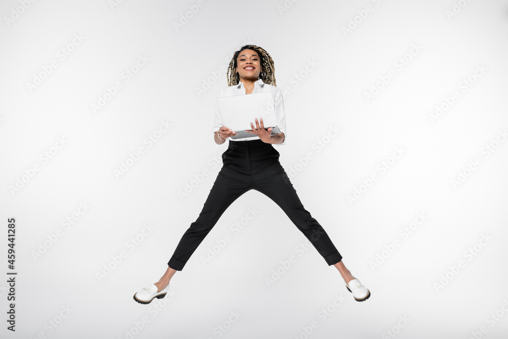 young african american businesswoman smiling at camera while levitating with laptop isolated on white young african american businesswoman smiling at camera while levitating with laptop isolated on white