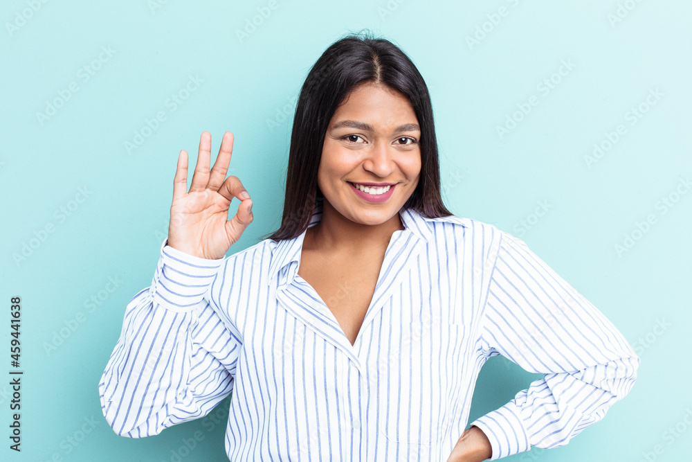 Young Venezuelan woman isolated on blue background winks an eye and holds an okay gesture with hand.