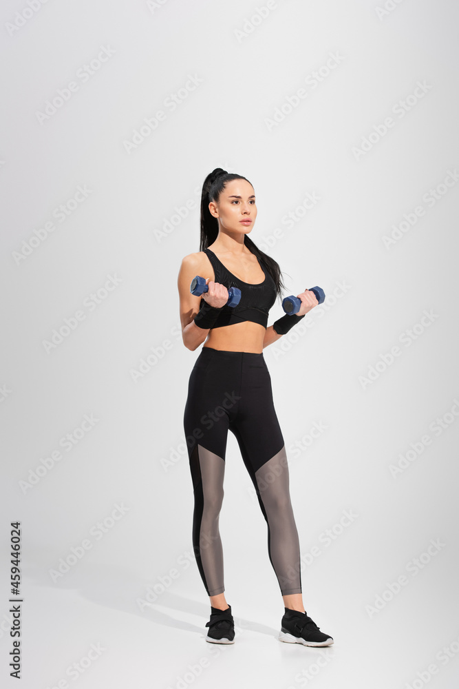 full length of strong young woman working out with dumbbells on grey full length of strong young woman working out with dumbbells on grey