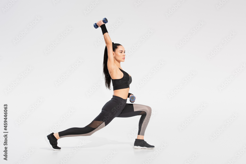full length of strong sportswoman in sportswear working out with dumbbells on grey