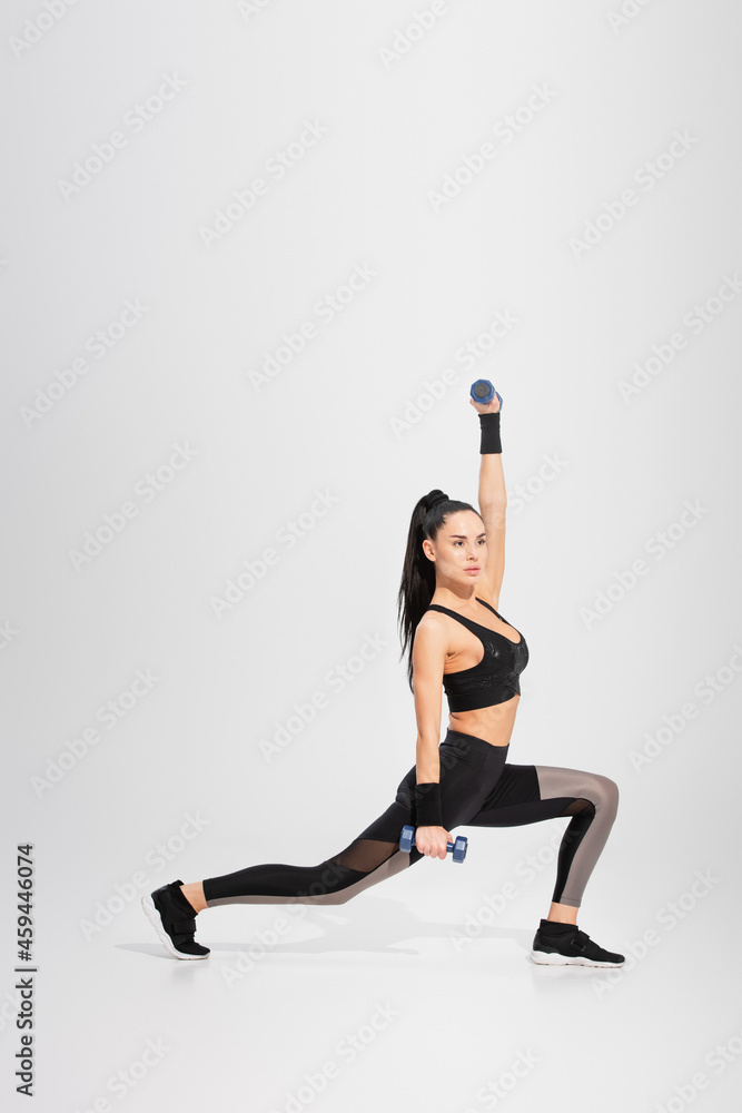 full length of strong young sportswoman in sportswear working out with dumbbells on grey