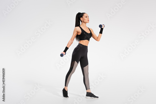 full length of strong woman working out with dumbbells on grey