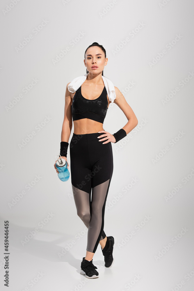 full length of young sportswoman with towel on shoulders holding sports bottle while posing with hand on hip on grey full length of young sportswoman with towel on shoulders holding sports bottle while posing with hand on hip on grey