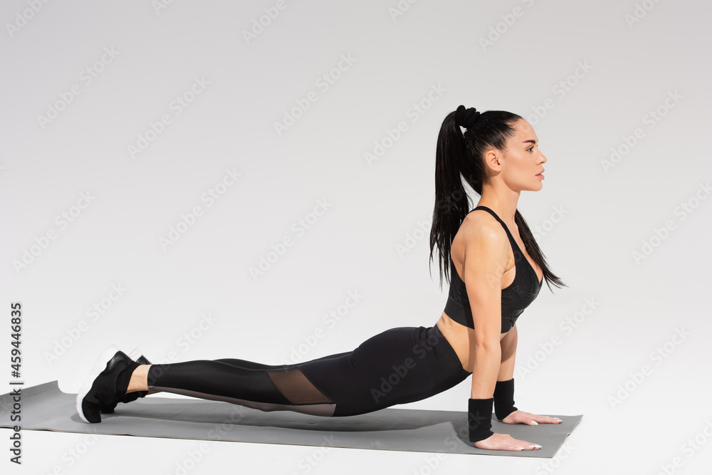 Fototapeta premium side view of young woman in black sportswear exercising on fitness mat on grey
