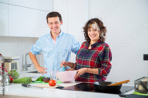 mujer joven sonriente bate huevos en un bol rosa con una varilla de cocina azul mientras rie con su pareja en una cocina blanca con verduras y vegetales