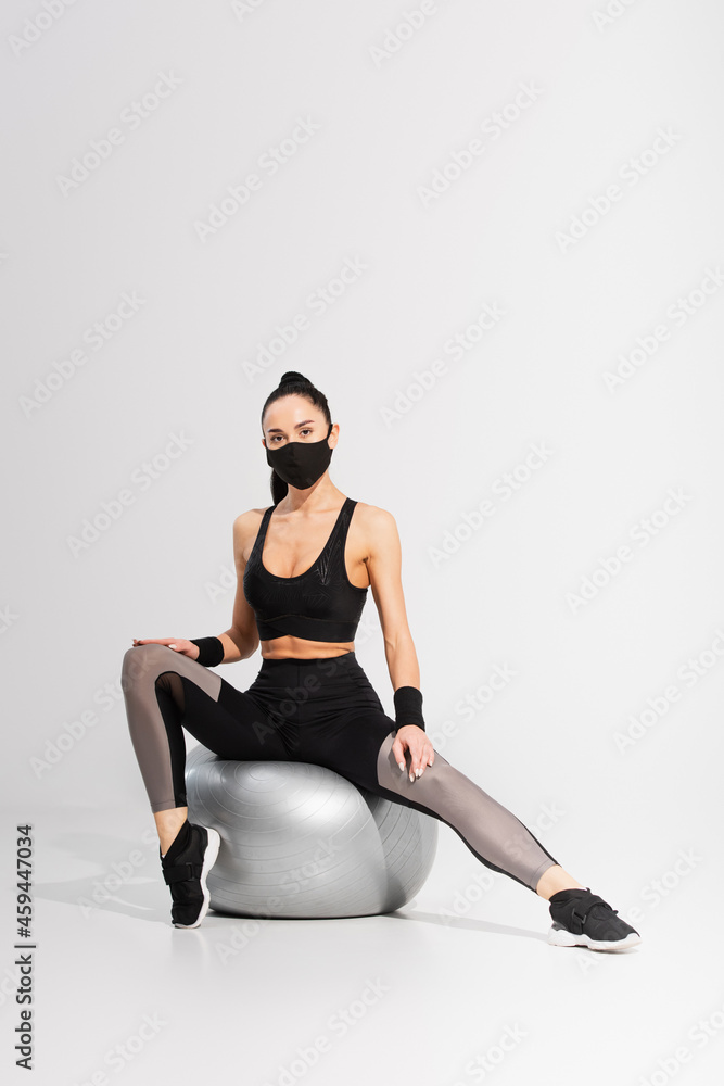 brunette sportswoman in black protective mask exercising on fitness ball on grey brunette sportswoman in black protective mask exercising on fitness ball on grey