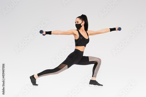 full length of young sportswoman in black protective mask exercising with dumbbells on grey