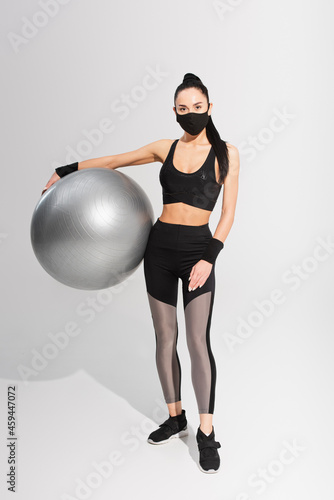 young sportswoman in sportswear and black protective mask holding fitness ball on grey