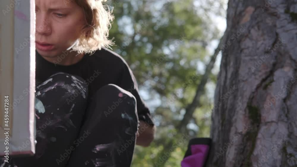 Modern stylish girl artist paints a picture in the forest, leaning a canvas on a tree. Focused on the process, frowns and creates. Young creative free woman artist paints in nature on canvas. Modern stylish girl artist paints a picture in the forest, leaning a canvas on a tree. Focused on the process, frowns and creates. Young creative free woman artist paints in nature on canvas.