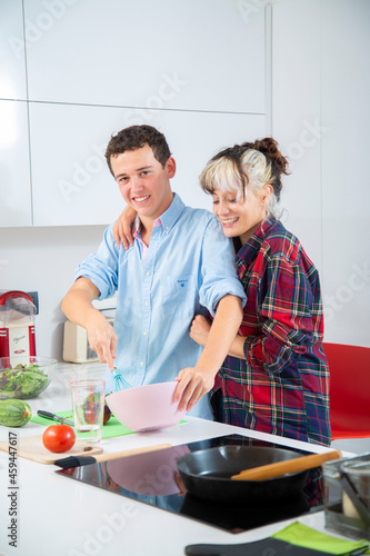 mujer joven sonriente bate huevos en un bol rosa con una varilla de cocina azul mientras rie con su pareja en una cocina blanca con verduras y vegetales
